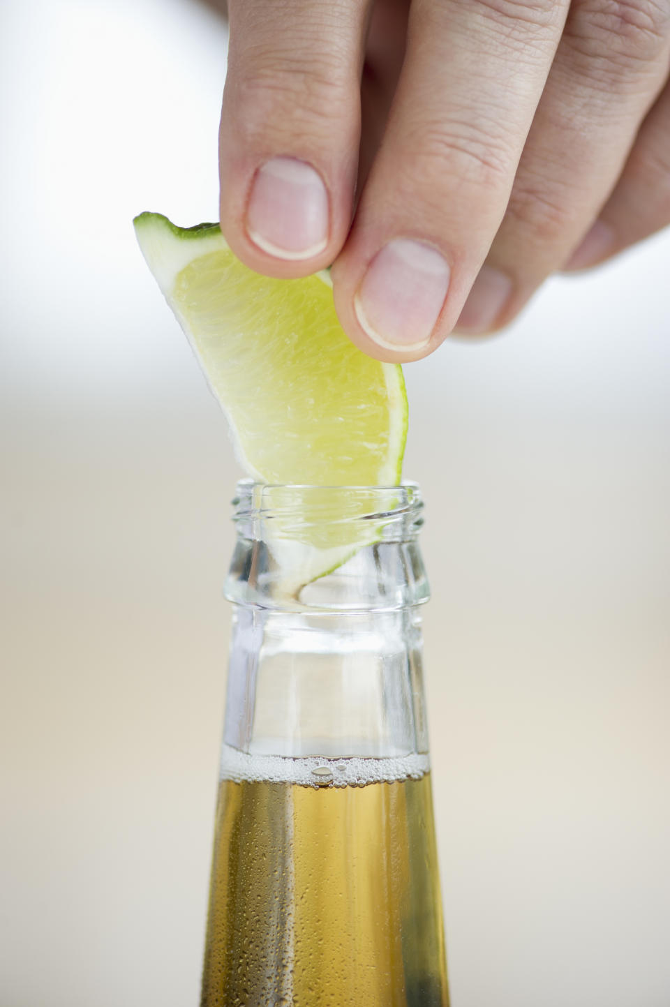 A hand is placing a lime wedge into the neck of a beer bottle. The focus is on the lime and the top of the bottle