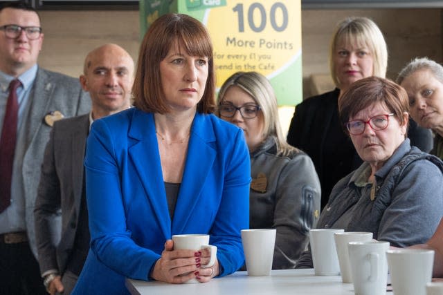 Rachel Reeves looking serious with her hands holding a white mug on a white table with people in the background