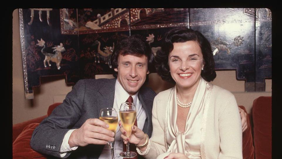 richard blum and dianne feinstein smile for a photo while sitting on a red couch and holding champagne flutes, he wears a gray suit with a white shirt and red tie, she wears a white sweater and blouse with pearls