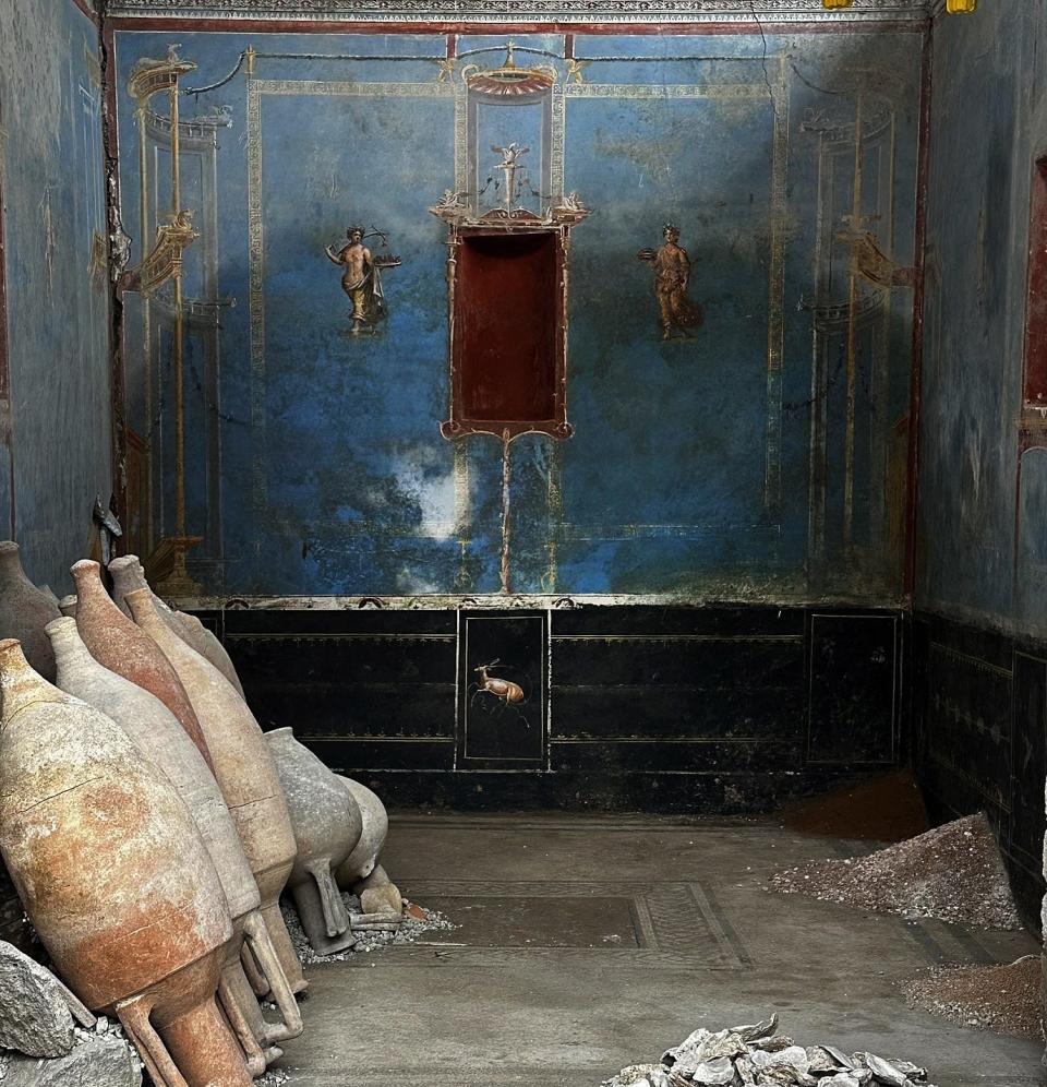 A blue-painted room in Pompeii with figures on the wall and several large jugs leaning against a wall