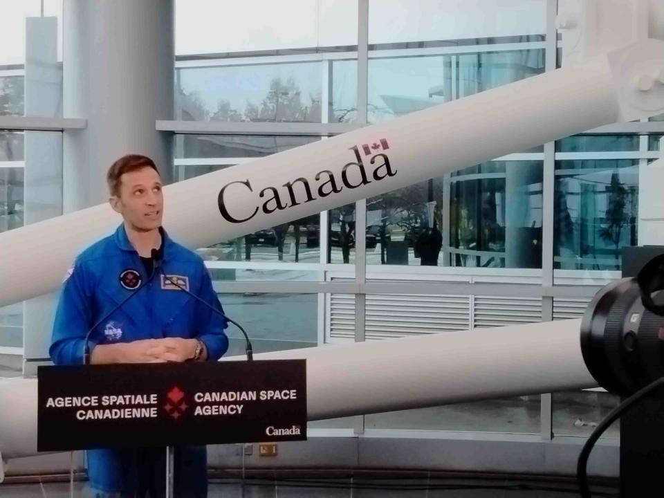 two people in blue flight suits speak at a podium in a large lobby lined with windows