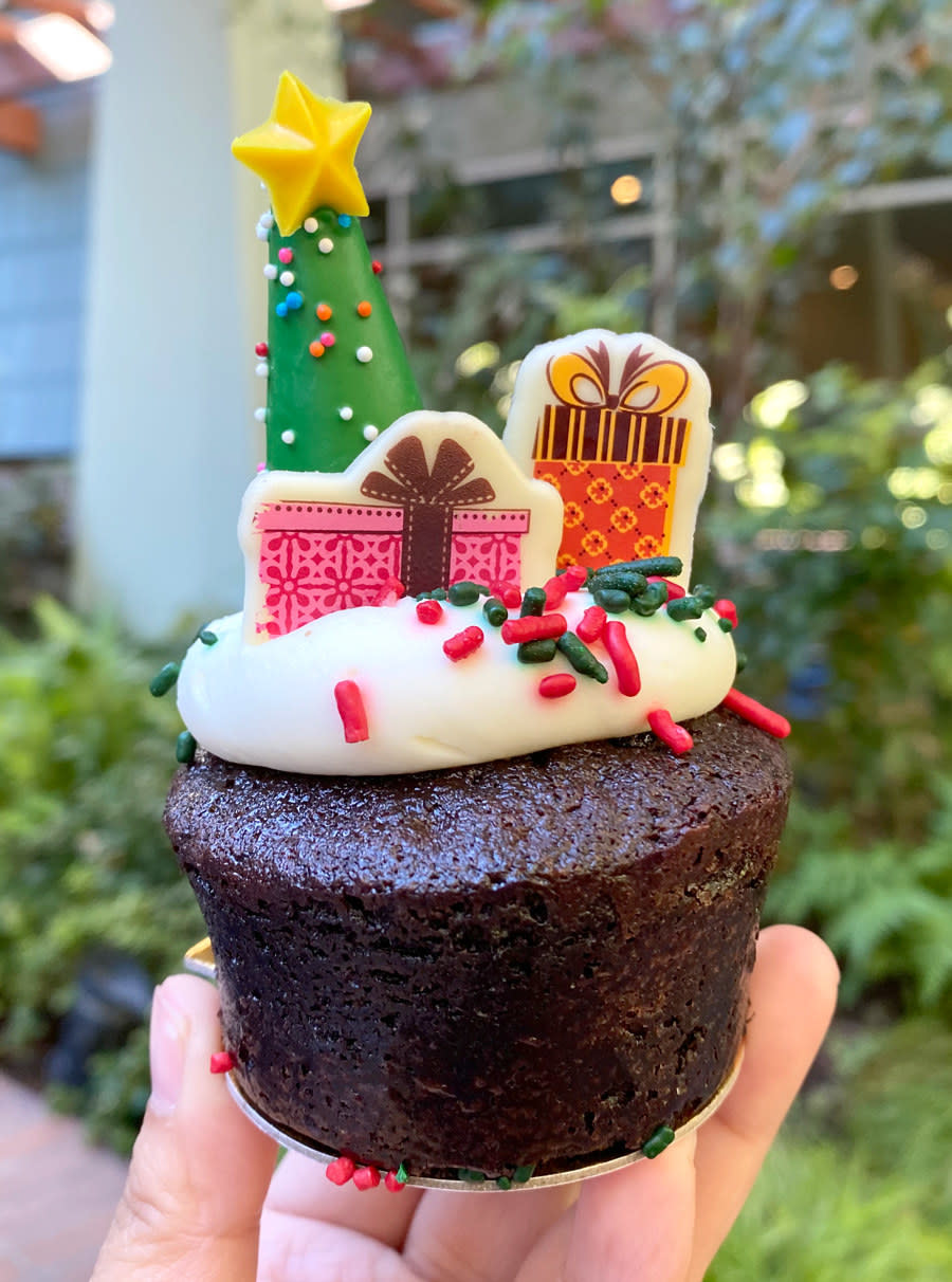 A cupcake sized brownie with frosting, sprinkles, and white chocolate shaped like a christmas tree and presents on top