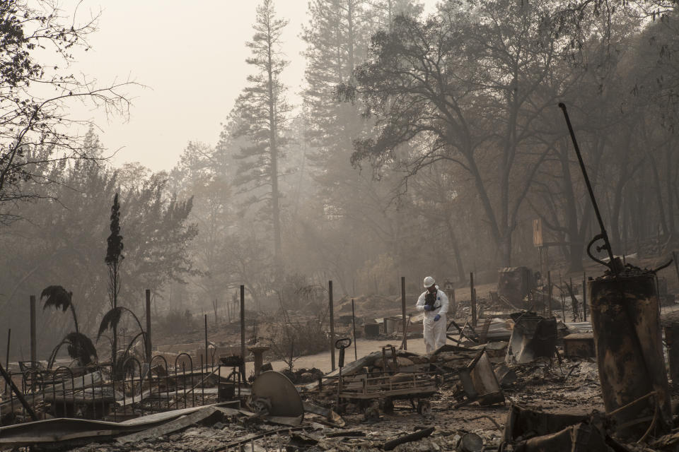 Person in white hazmat-type coveralls and white helmet stands looking down near the burned wreckage of where a home apparently once stood.