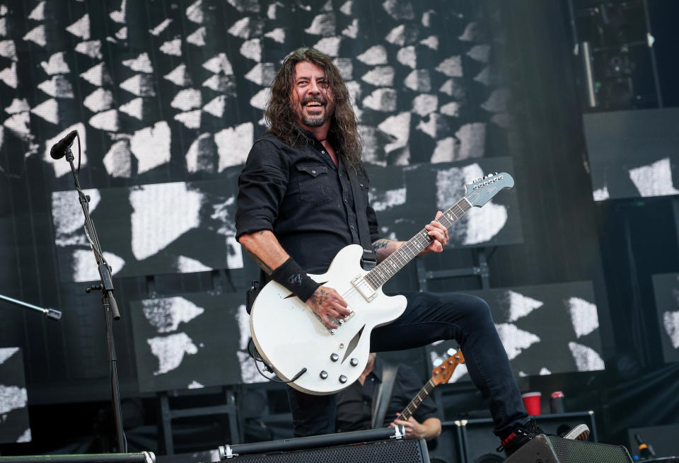 Dave Grohl playing an electric guitar on stage, smiling, wearing a dark long-sleeve shirt and dark jeans. There is a microphone stand and a large screen in the background