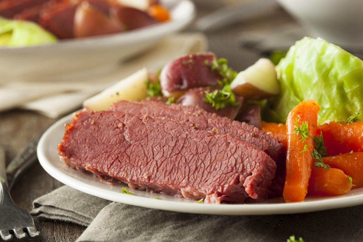 Instant pot corned beef with potatoes and vegetables on a white plate on a grey napkin with a fork with a blurred background of food