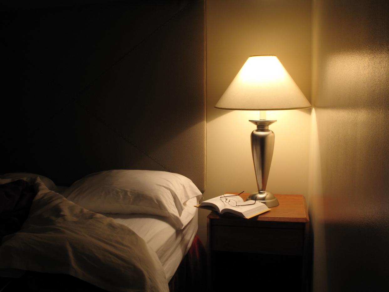 Night scene in hotel room, nightstand with lamp, bed turned down, opened book and reading glasses. 1st in series. Great wall reflection and texture.