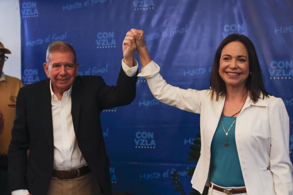 A man in a white shirt and dark jacket holds the hand of a smiling woman in a blue top and white jacket, right