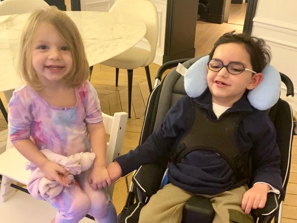 Eva Vauclare, 3, sits on a white stool and holds hands with her older brother, Leo, as he sits in a moulded seat that supports his head and body,