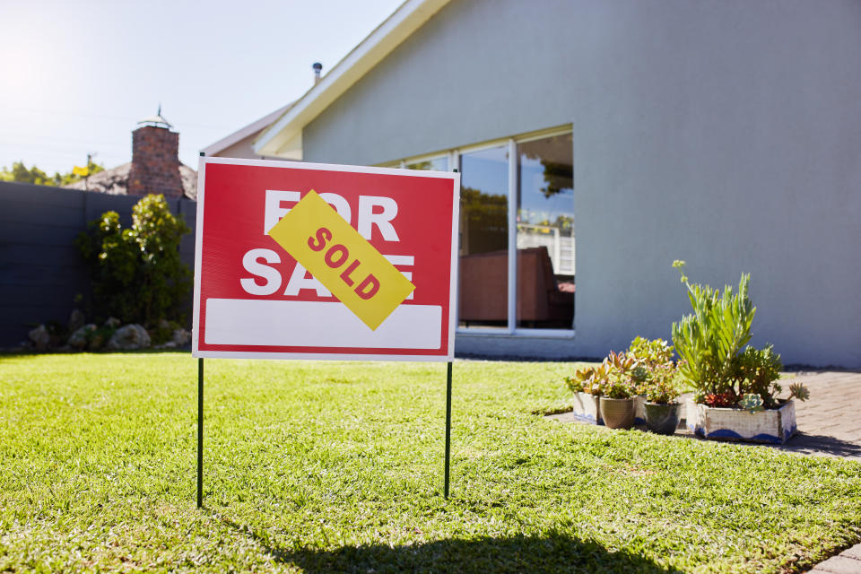 A "For Sale" sign with a "Sold" sticker is displayed in front of a modern house with a well-maintained lawn and garden