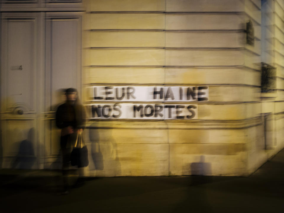 In this Nov. 8 2019 photo, Sarah stands next to slogan reading "Their hate, our dead" in Paris. Under cover of night, activists have glued slogans to the walls of buildings to draw attention to domestic violence, a problem French President Emmanuel Macron has called "France's shame." (AP Photo/Kamil Zihnioglu)