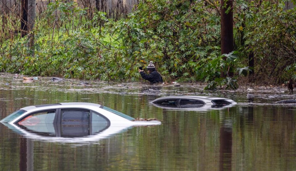 The roofs of two cars are visible amid high water. In the background, a man in a coat and beanie moves through the water.