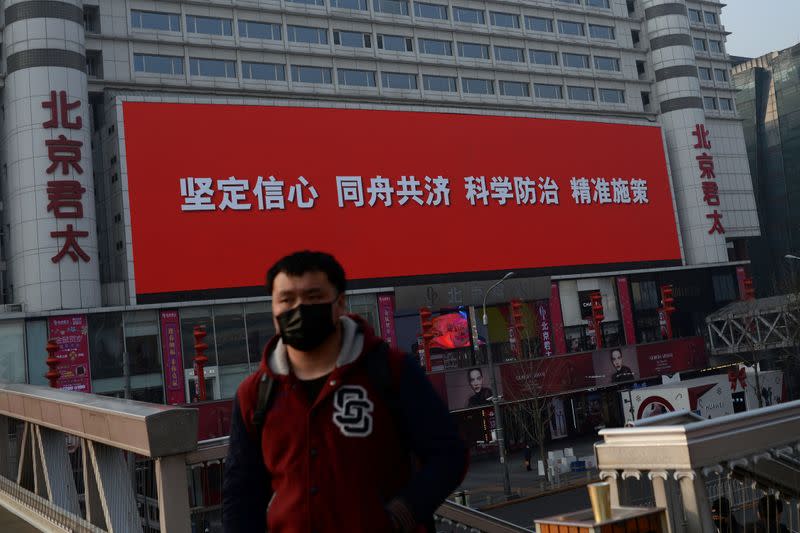 Man wearing a face mask stands near a giant screen showing a propaganda slogan to fight against the novel coronavirus in Beijing
