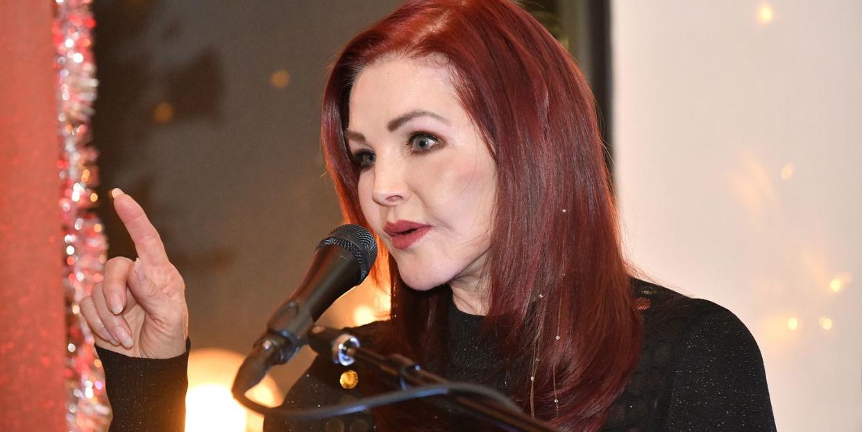 priscilla presley speaks into a microphone and holds her right hand up while pointing her index finger, she is wearing a black long sleeve shirt and makeup