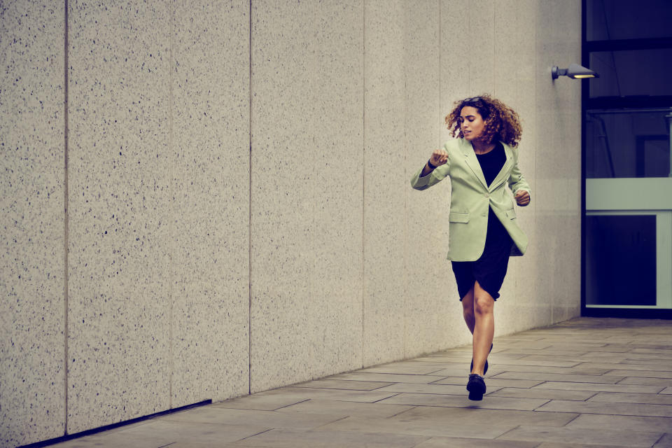 A woman in a professional outfit is walking briskly outdoors beside a modern building. She appears focused and determined