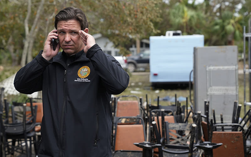 Florida Gov. Ron DeSantis talks on the phone as he stands outside a storm-damaged restaurant. Stacks of chairs and kitchens supplies are seen behind him.