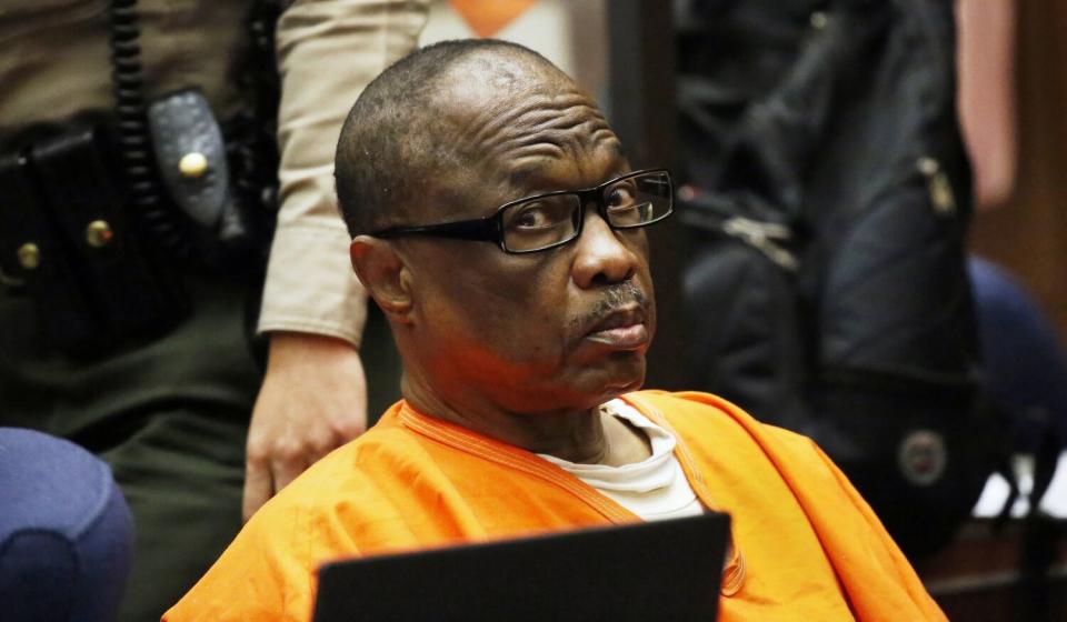 A seated man wearing glasses and orange prison garb