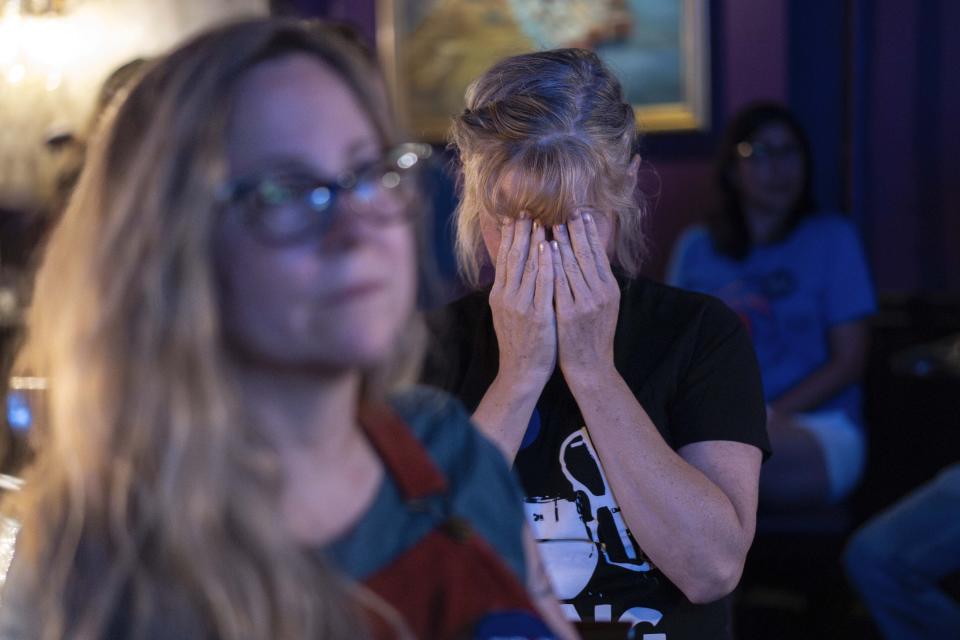 Two people in a room; one woman in the foreground wears glasses, while another places her hands over her face, appearing distressed or emotional