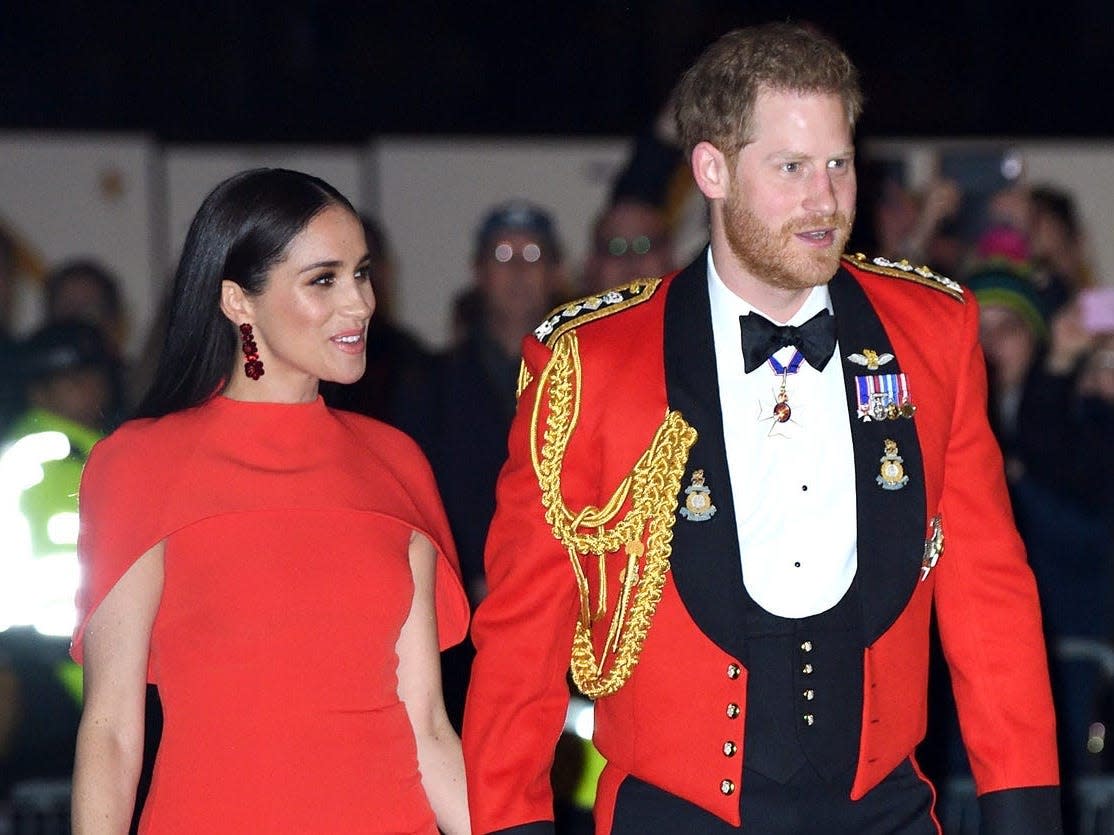 Meghan in a red cape sleeve dress and Harry in a red military jacket with badges, ropes, and a bow tie.