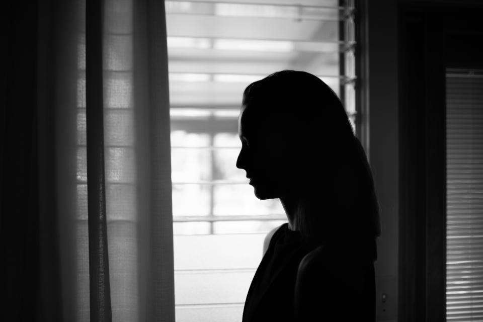 Silhouette of a person standing next to a window, looking outside through partially open blinds