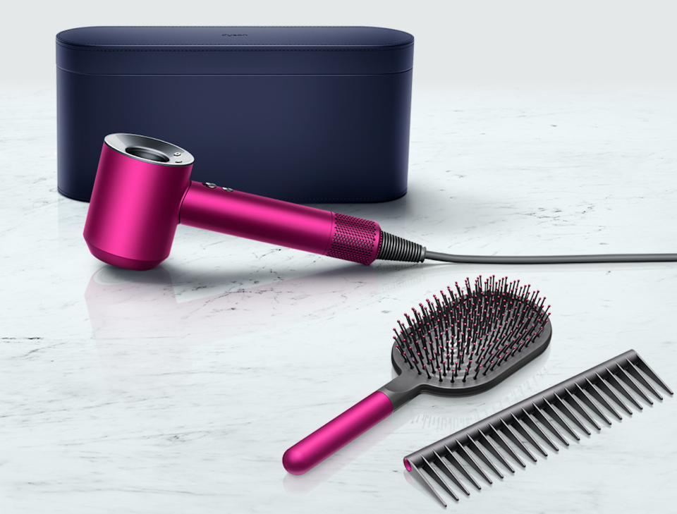 A dark blue oval case sits on a marble tile behind a hot pink Dyson dryer, and matching brush and comb.