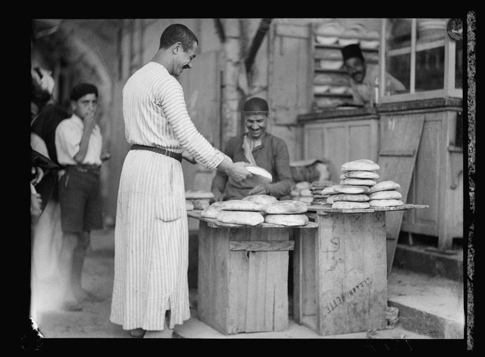 A black and white photo of a Palestinian baker selling bread to a customer.