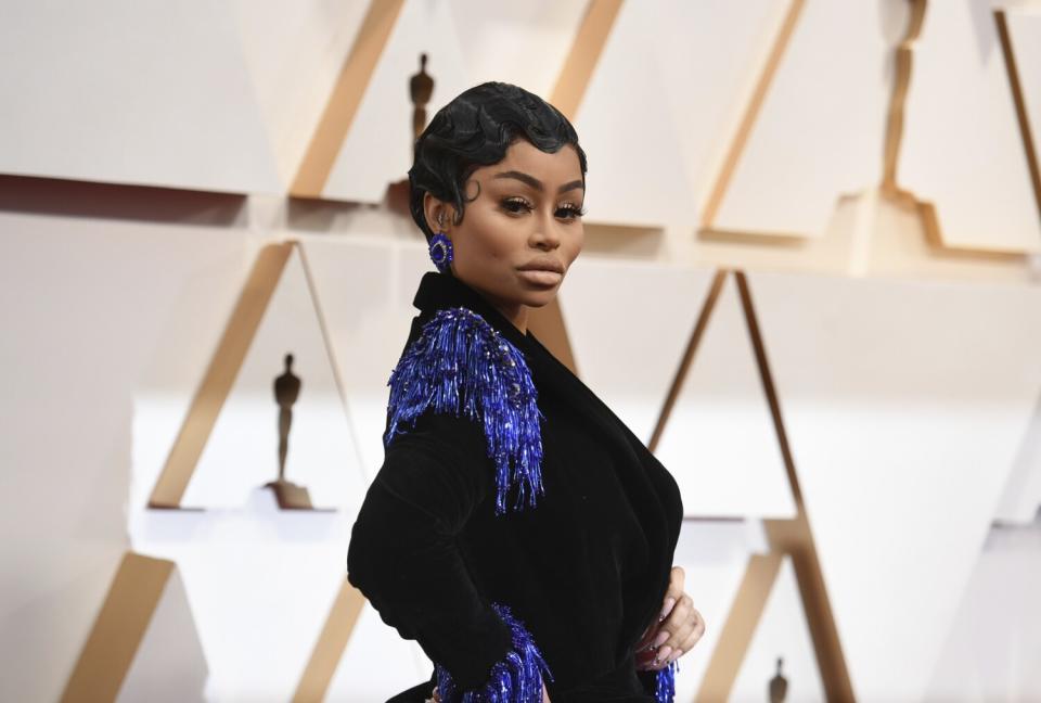 A woman wearing a black dress with purple accents poses in front of Oscar statuettes