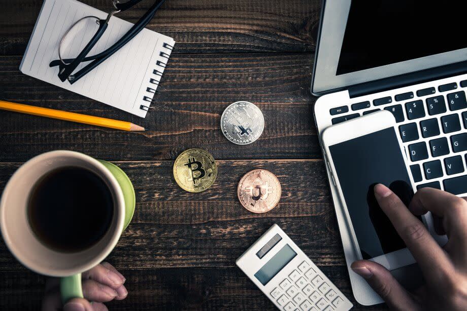 Bitcoin-Münzen nebst einer Kaffeetasse, einer Brille, einem Notizblock, einem Taschenrechner, einem Smartphone und einem Laptop