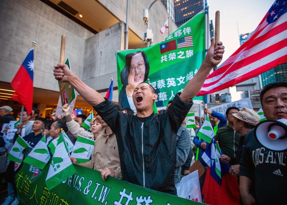 A young man stretches out his arms while chanting during a protest. People hold Taiwanese and American flags.