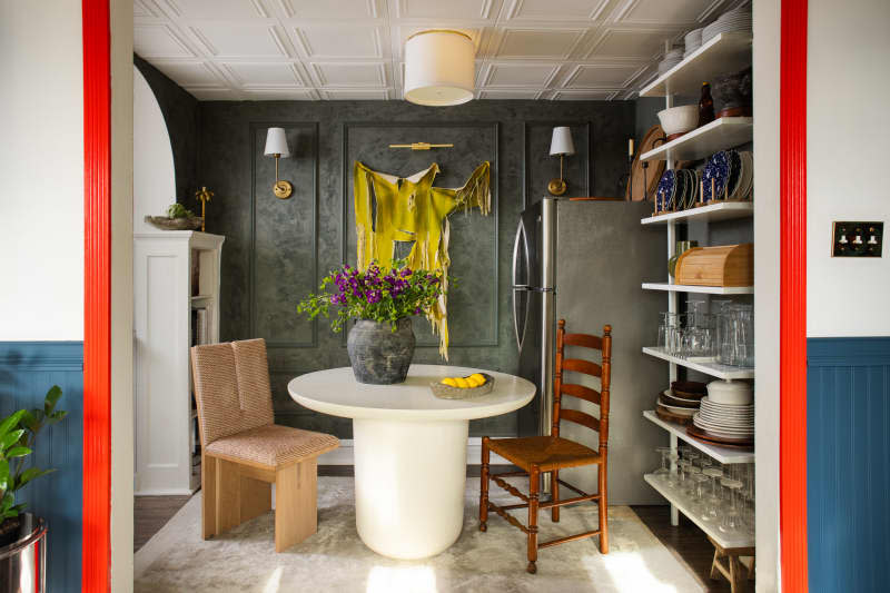 Dining room with gray back wall and white round table. Shelves with dishes and glassware on the right