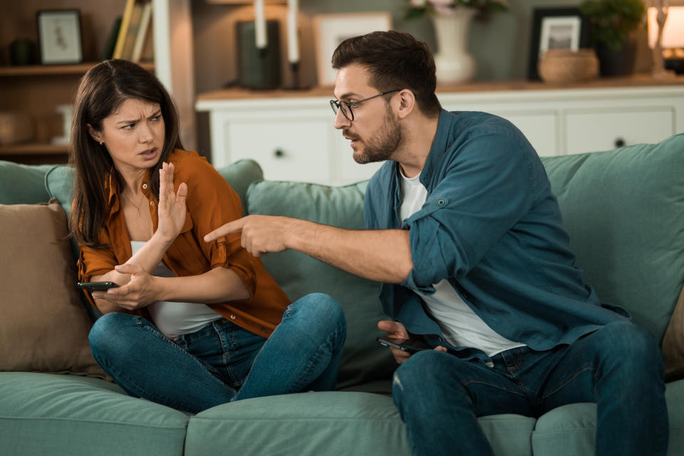 A woman and man sitting on a couch arguing. The woman raises her hand, while the man points at her. Both hold smartphones