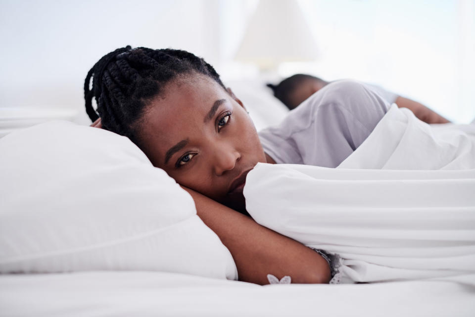 A woman looking concerned in bed while her husband sleeps on the other side