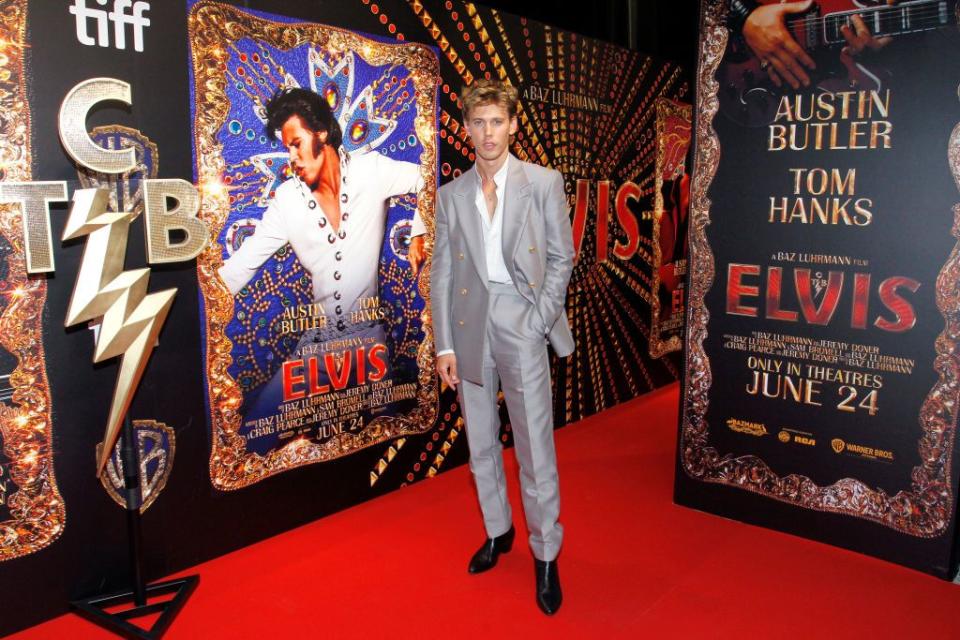 austin butler wearing a silver suit and standing next to a film poster showing him in costume as elvis presley