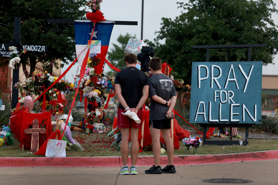 People stand in front of a makeshift memorial that includes flowers, flags and a sign that reads: Pray for Allen.