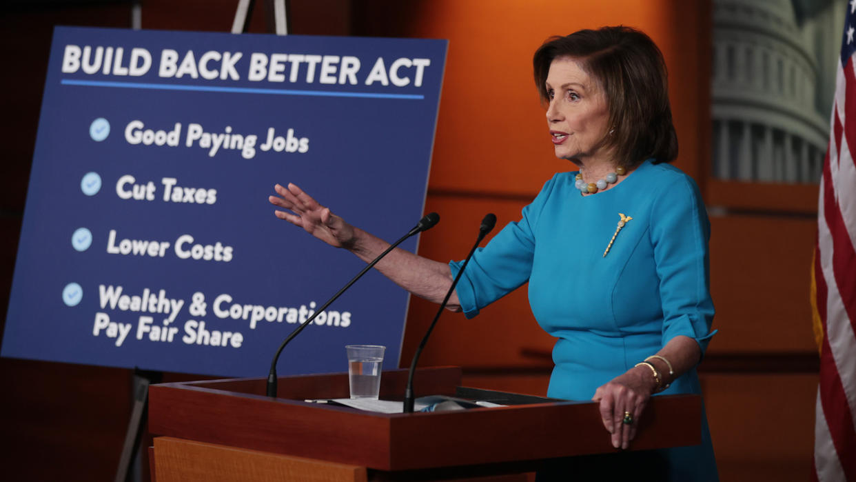 House Speaker Nancy Pelosi stands before a signboard labeled 