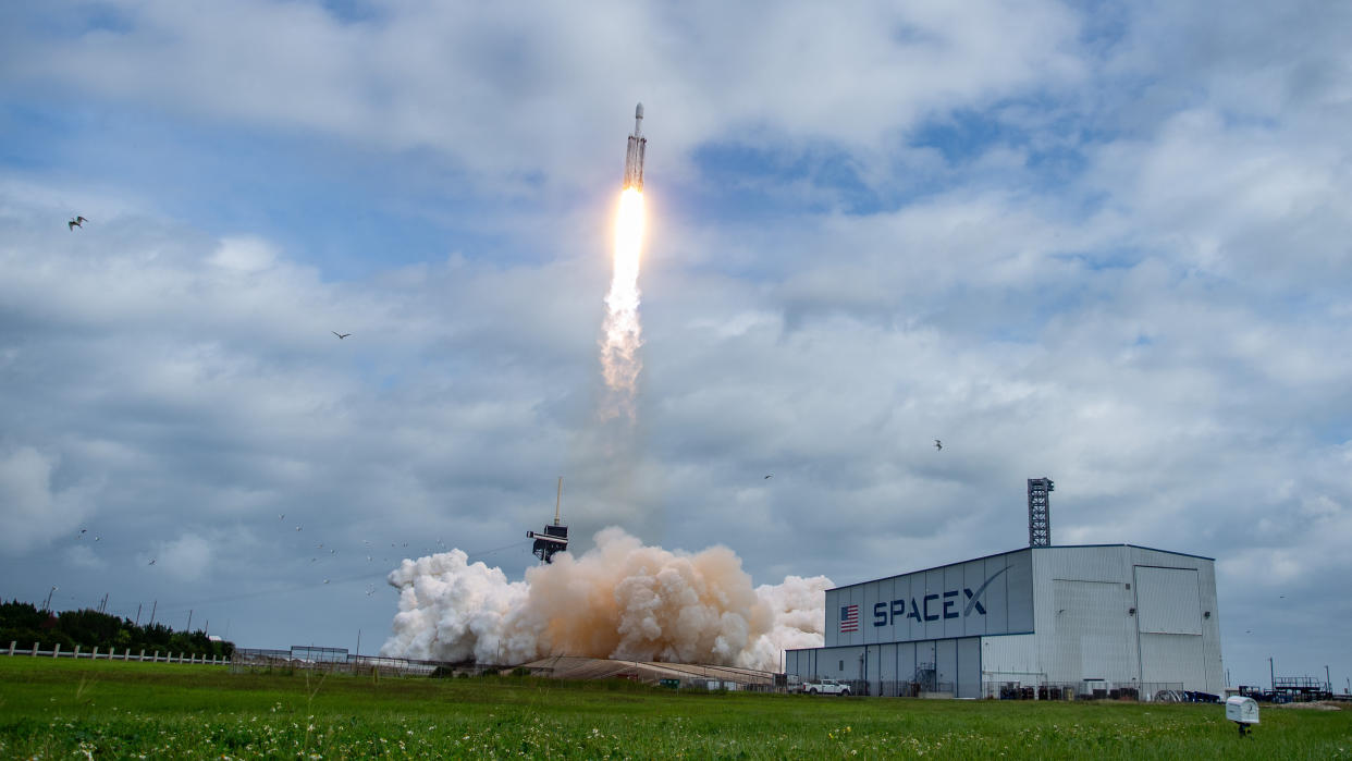  A triple-core rocket blasts off under a sunny, partly cloudy blue sky. smoke expands below next to an obscured launch tower, near a long white hanger with SPACEX written on the side. To the left, a bird flies away. 