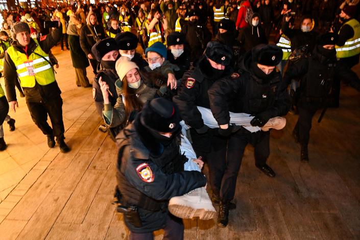 A group of seven Russian police officers surround a woman in a wool hat, two holding her left leg, and one her other leg, as other civilians stand in the background.
