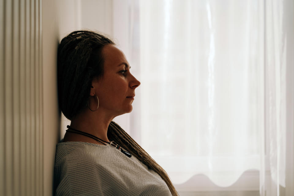 A woman with long braided hair sits pensively against a wall, looking ahead. She wears hoop earrings and a light top. The background features softly lit curtains