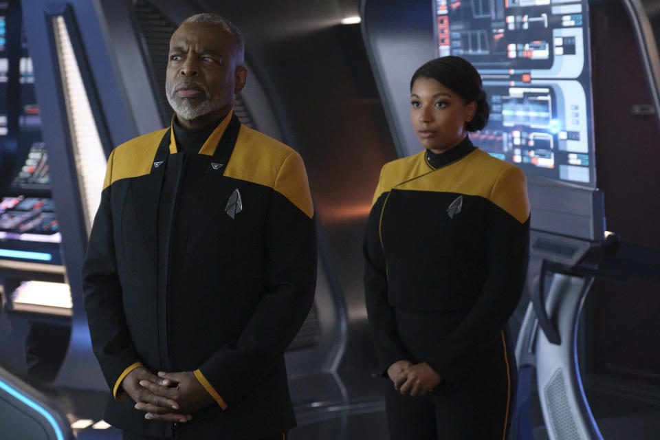 Two Star Trek characters in uniform stand on the bridge of a starship, looking serious
