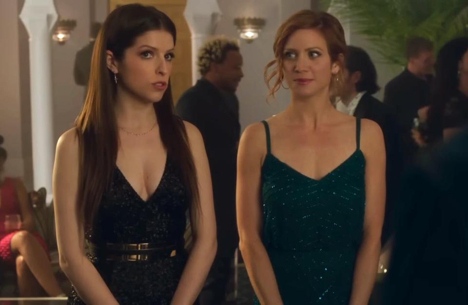 Anna Kendrick and Brittany Snow are dressed in elegant evening gowns, standing at a social event indoors, possibly a party or reception