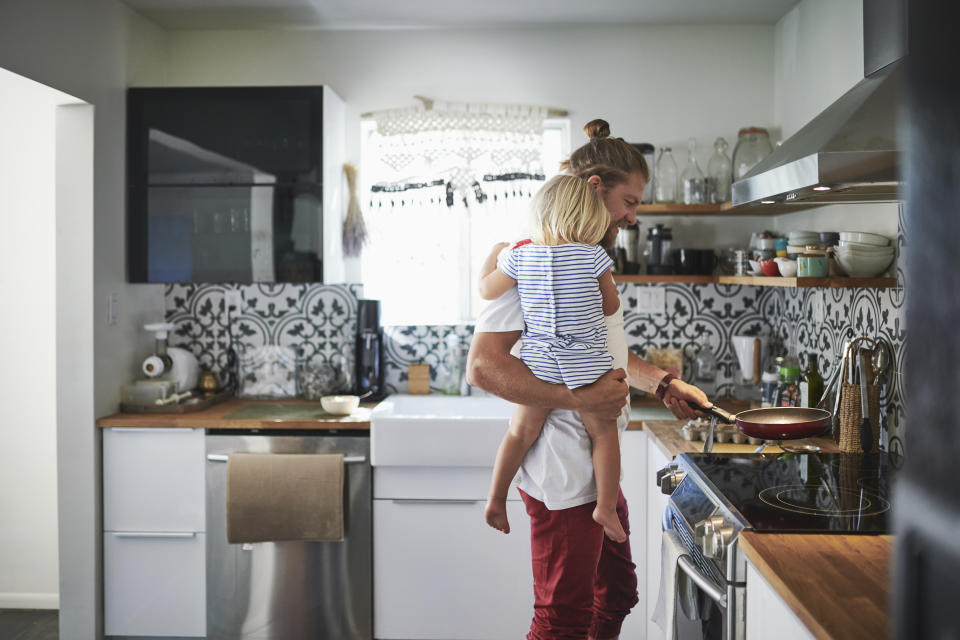 A man holds a toddler while cooking in the kitchen to represent a father taking paid parental leave.