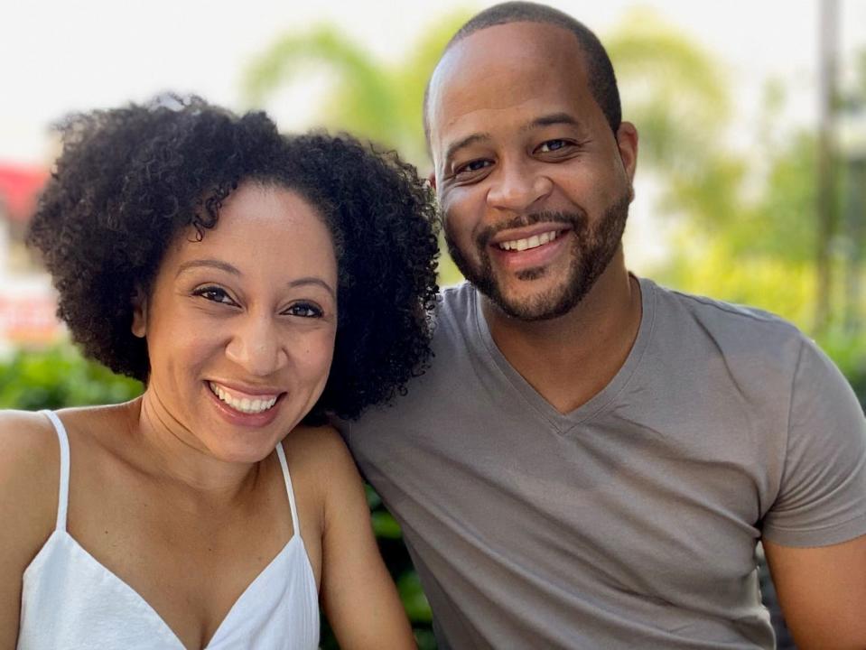 A photo of Stephanie Claytor and her husband, Corey, sitting at a restaurant. Stephanie has chin-length curly black hair, brown eyes, and wears a white strappy dress with a tie in the front. Her husband has a shaved fade hair cut, closely cropped mustache and beard. He wears a grey V-neck t-shirt. They lean close together, smiling.