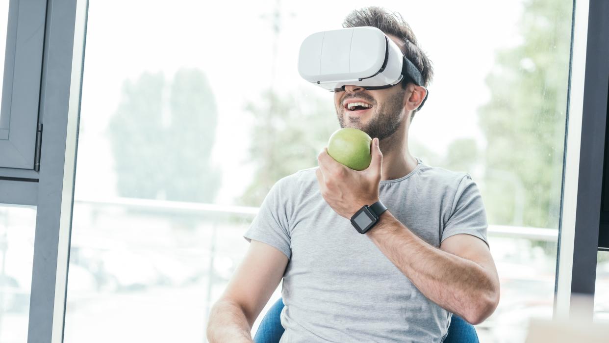  smiling young man holding green apple and using virtual reality headset 