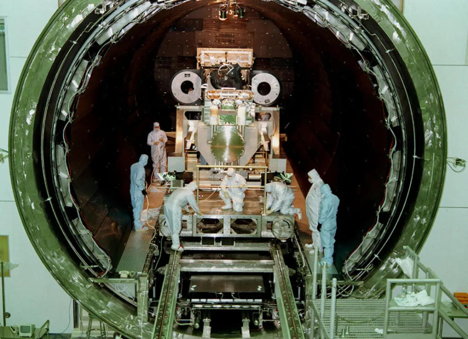 Clean-suited technicians attend to a large piece of machinery in a circular doorway that spans several metres.