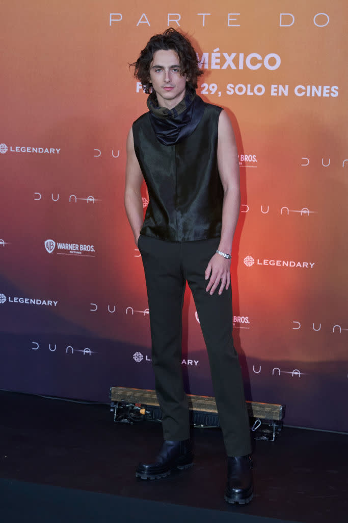 Timothée at event wearing sleeveless top and trousers, standing in front of a promotional backdrop