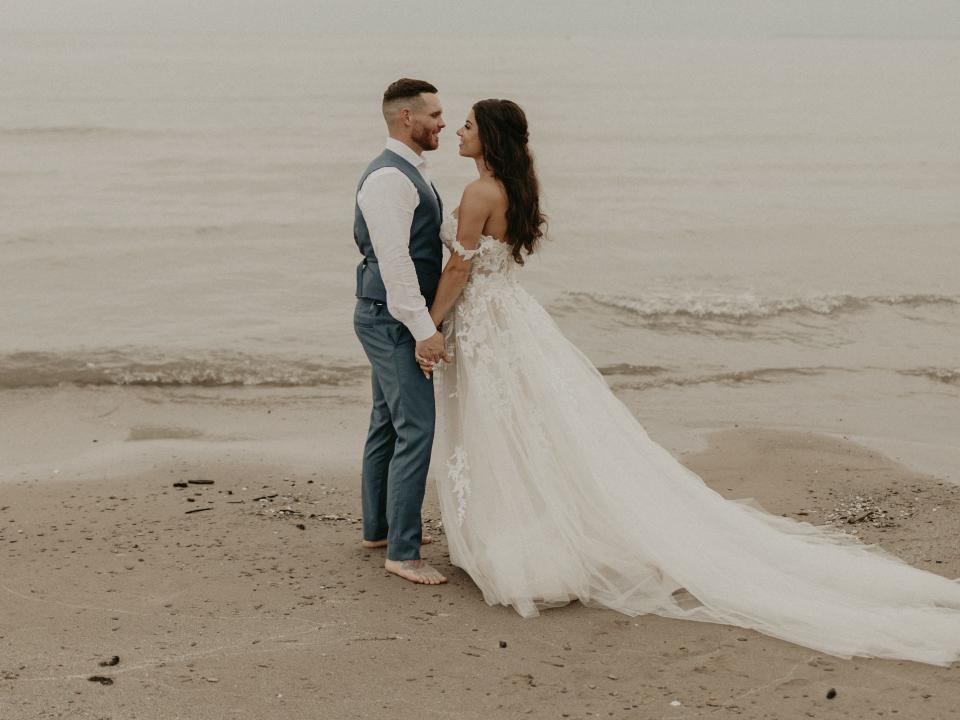 A bride and groom hold hands and gaze at each other in front of the ocean.