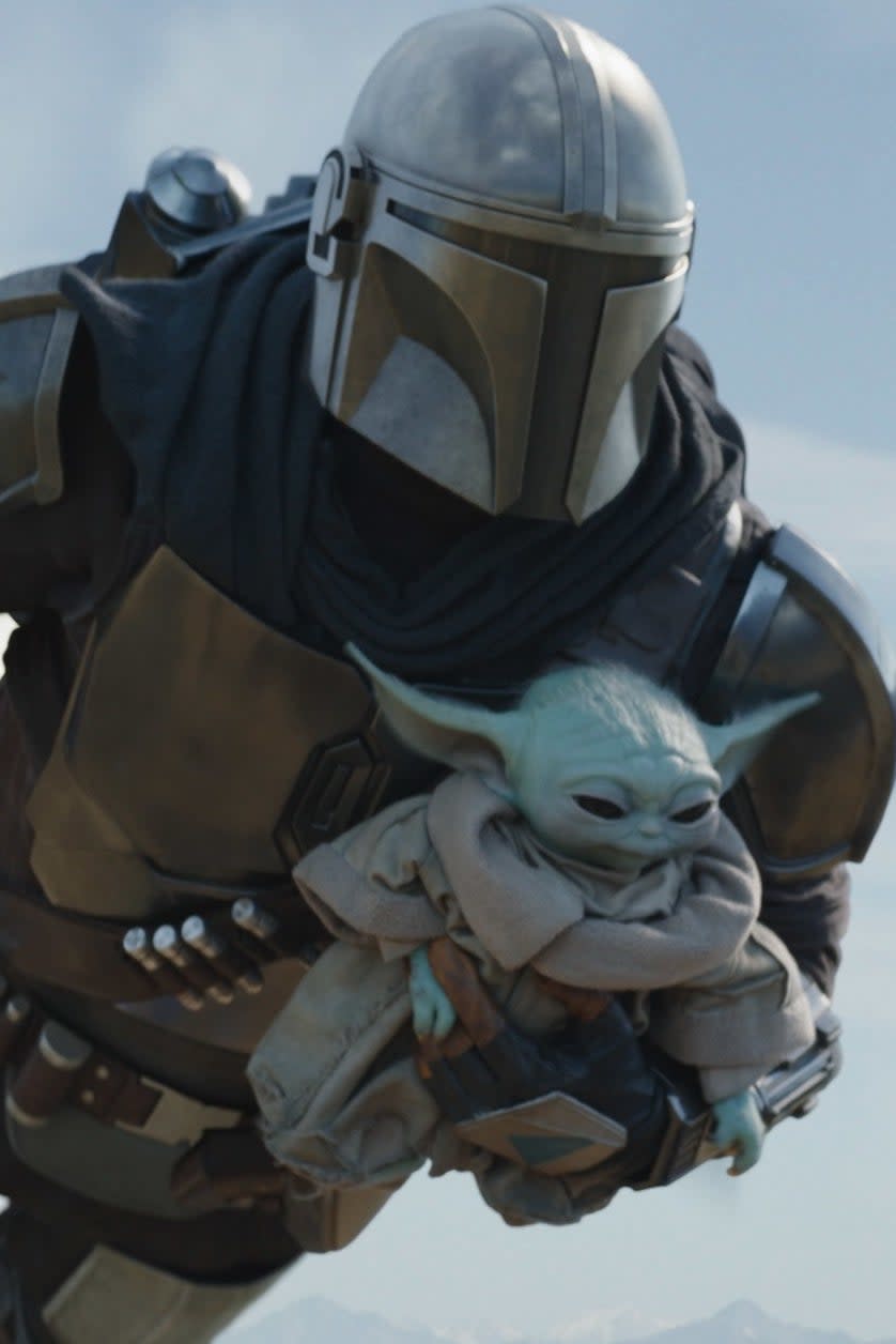 The Mandalorian, in armor, holds Grogu while flying through the sky