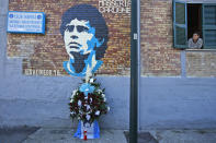 A woman looks out of a window as flowers commemorating soccer legend Diego Maradona are placed beneath a mural depicting him, in Naples, southern Italy, Thursday, Nov. 26, 2020. Maradona died Wednesday, Nov. 25, 2020 in Buenos Aires. (AP Photo/Alessandra Tarantino)