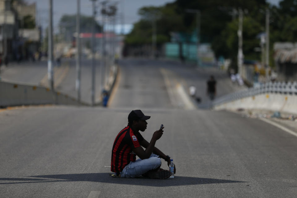 A man using his cell phone sits in the shade of a street sign in the middle of a normally busy main road that was devoid of drivers, as protesters calling for the resignation of President Jovenel Moïse gathered at a bridge in the Delmar area of Port-au-Prince, Haiti, Thursday, Oct. 17, 2019. Haiti's embattled president was forced on Thursday to hold a private ceremony amid heavy security for what is usually a public celebration of one of the country's founding fathers, revolution leader Jean-Jacques Dessalines. (AP Photo/Rebecca Blackwell)