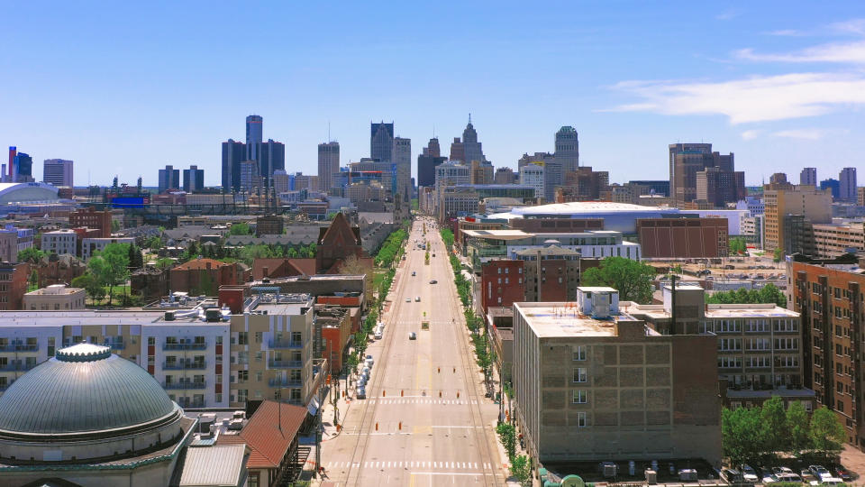 An aerial view of Detroit, showing the wide, empty Woodward Avenue and high-rise buildings in the distance.