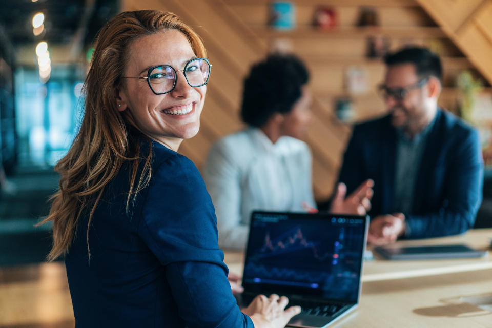 Portrait of crypto trader at the office. Successful female trader in formalwear working in office, looking at camera in front of laptop screen, checking global currency index on fund exchange. Trading stock or cryptocurrencies. Trading app on screen.